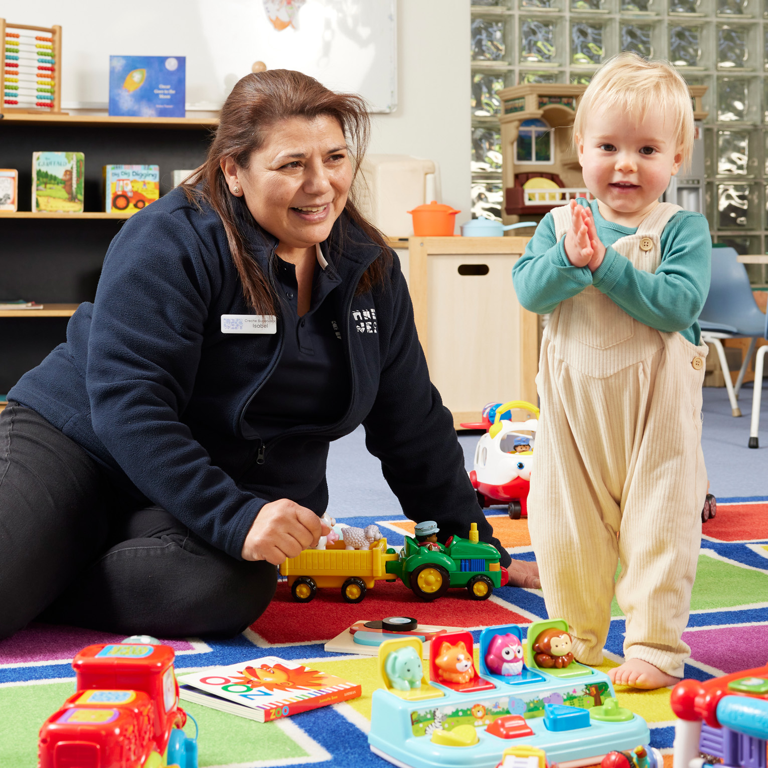 Creche assistant sitting on the floor on a colourful mat surrounded by toys with a young child standing and clapping their hands